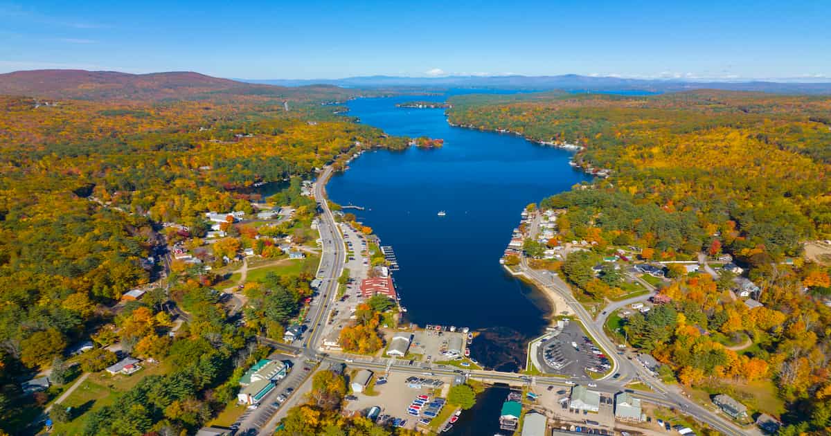 Aerial view of a lake surrounded by trees with autumn foliage, small towns, roads, and docks along the shoreline under a clear blue sky.
