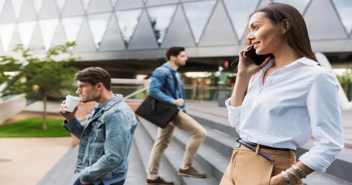 Three young adults outside an office building: a woman in front talking on her phone, a man in a denim jacket drinking coffee, and another man walking up the steps with a bag. Modern glass building in the background.