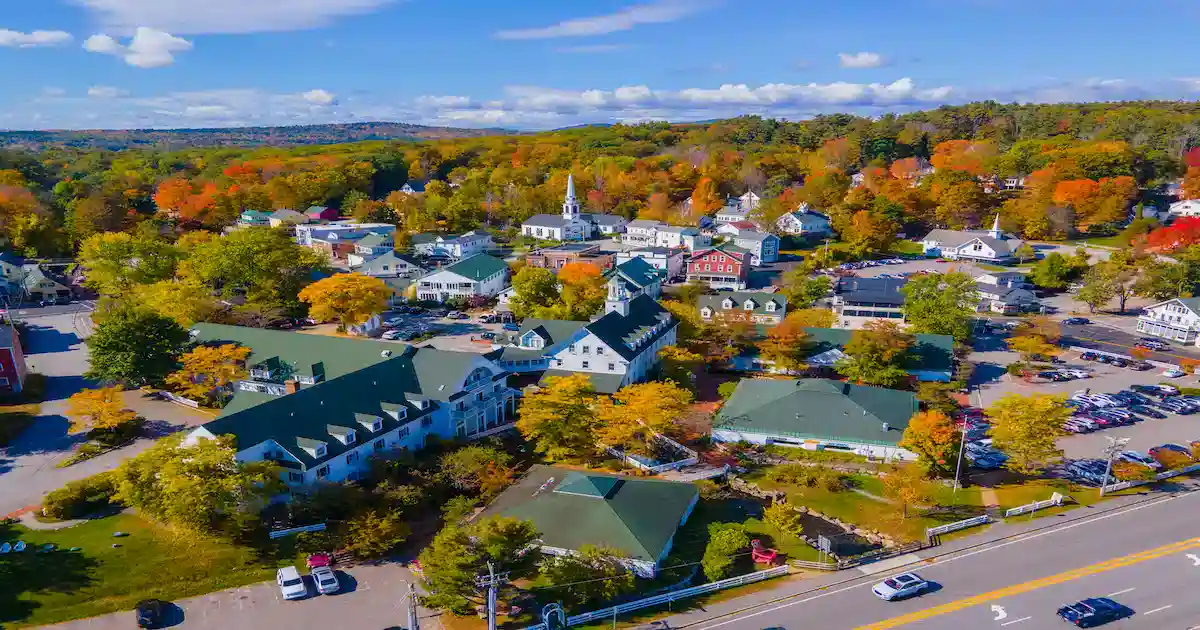 Aerial view of a small town with green-roofed buildings, colorful autumn trees, a white church steeple, parked cars, and a road in the foreground under a blue sky with scattered clouds.