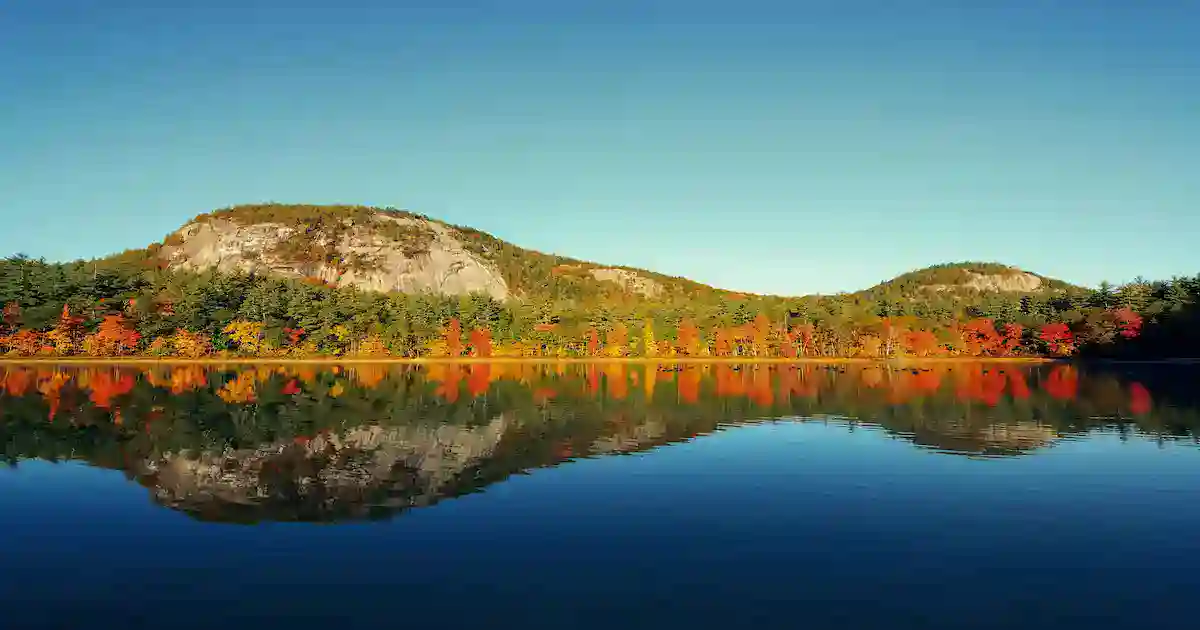 A calm lake reflects forested hills with vibrant autumn foliage in shades of red, orange, and yellow under a clear blue sky.