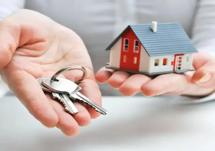 A person holding a set of keys in one hand and a small model house in the other, symbolizing buying, selling, or renting a home.