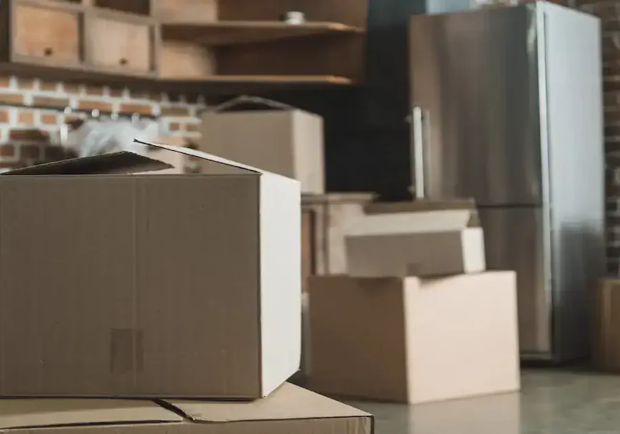 Several cardboard boxes, some open and some closed, are scattered around a kitchen with exposed brick walls, wooden shelves, and a stainless steel refrigerator, suggesting a moving or unpacking scene.