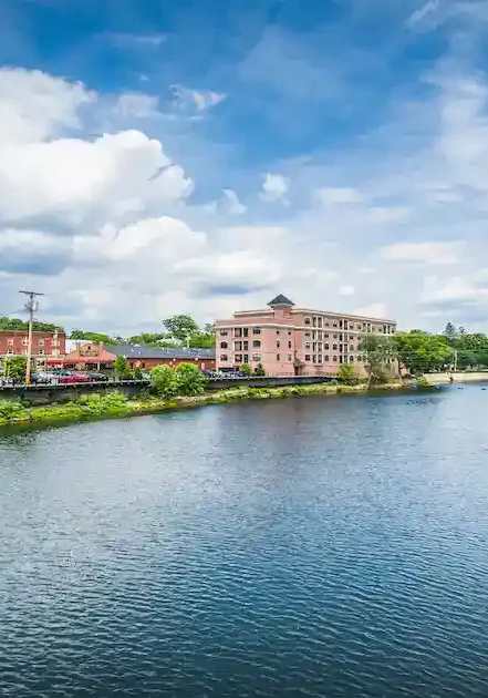 A calm river flows between brick buildings under a partly cloudy sky, with green trees and bushes lining the waters edge and a few distant cars visible on the left.