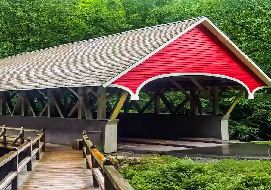 A red covered wooden bridge with a peaked roof crosses over a small stream, surrounded by lush green trees and foliage. A wooden walkway leads up to the bridge.