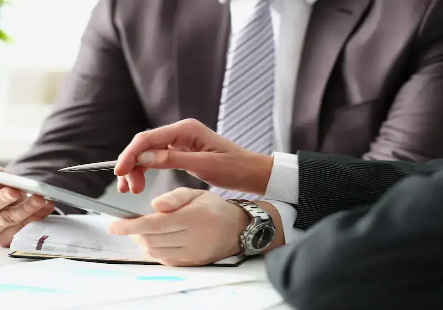 Two men in business suits are sitting at a desk, reviewing information on a tablet. One man is holding a pen and pointing at the tablet, while the other holds the device. A notebook and paperwork are visible on the desk.