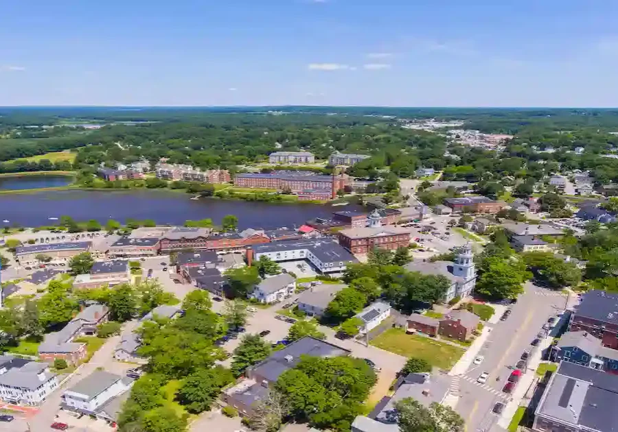 Aerial view of a small town with tree-lined streets, houses, buildings, a church, and a lake in the background under a clear blue sky. Lush greenery surrounds the town, with distant forests and fields visible.
