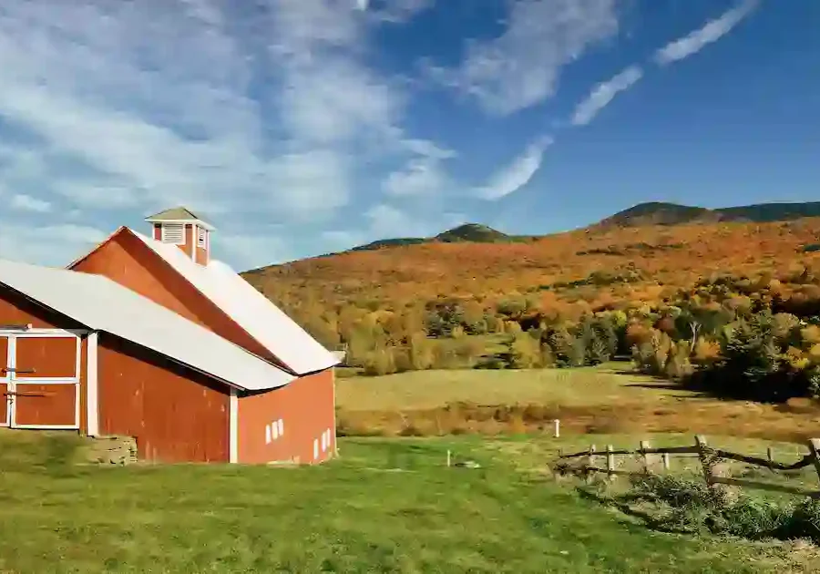 A red barn with a white roof sits on green grass in front of rolling hills covered in autumn trees under a blue sky with wispy clouds.