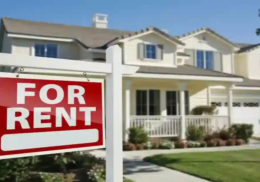 A large red and white For Rent sign is displayed in front of a modern, two-story house with a porch, green lawn, and a well-maintained garden.
