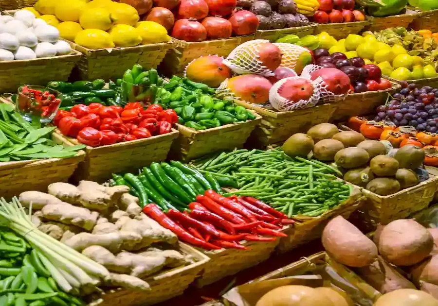 An assortment of fresh fruits and vegetables displayed in baskets at a market, including tomatoes, lemons, mangoes, grapes, ginger, sweet potatoes, green beans, chilies, and bell peppers, arranged in colorful rows.