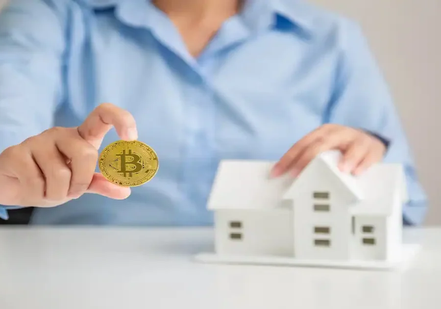 A person in a blue shirt holds a gold Bitcoin coin in one hand and touches a small white model house with the other, symbolizing cryptocurrency and real estate investment.