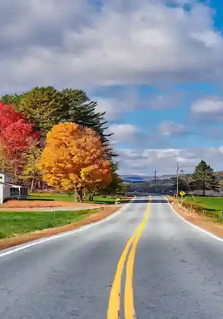 A paved country road with yellow center lines curves through green fields and trees with vibrant autumn foliage. A white house sits on the left, and the sky is partly cloudy with patches of blue.