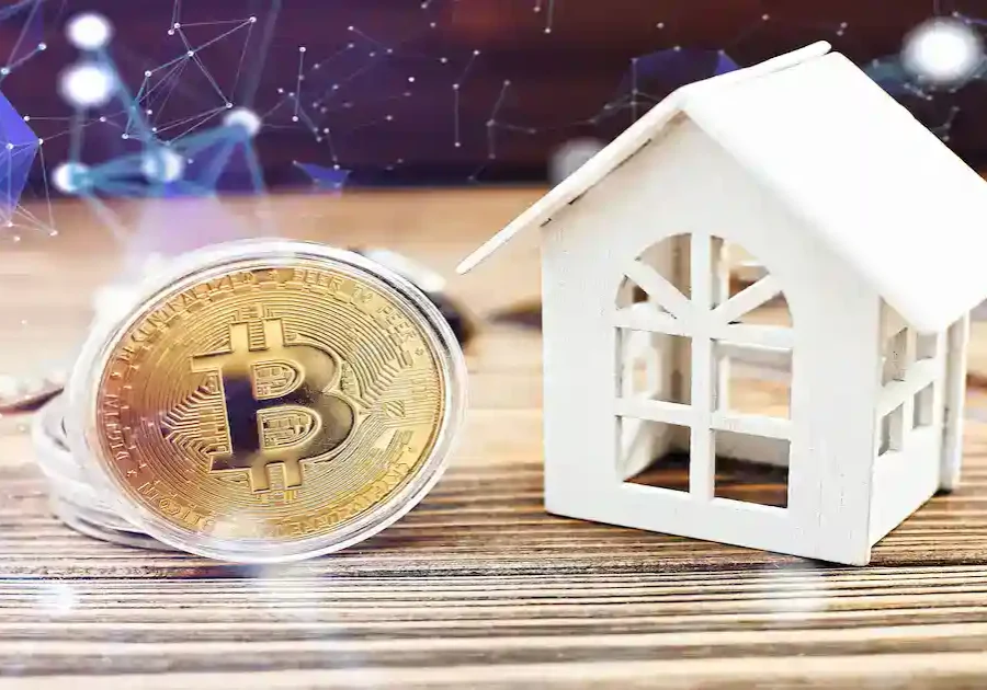 A gold Bitcoin coin stands upright next to a small white model house on a wooden surface, symbolizing the connection between cryptocurrency and real estate investment. Digital network graphics are overlaid in the background.