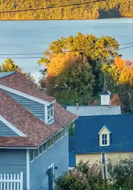 A residential street slopes toward a blue lake, surrounded by trees with green and autumn-colored leaves. Houses with varied roofs line the street, and forested hills rise in the background.