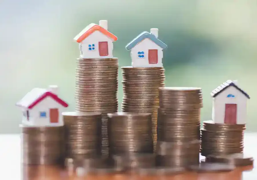 Four miniature houses sit atop stacks of coins of varying heights, symbolizing different property values or real estate investment growth, with a blurred green background.
