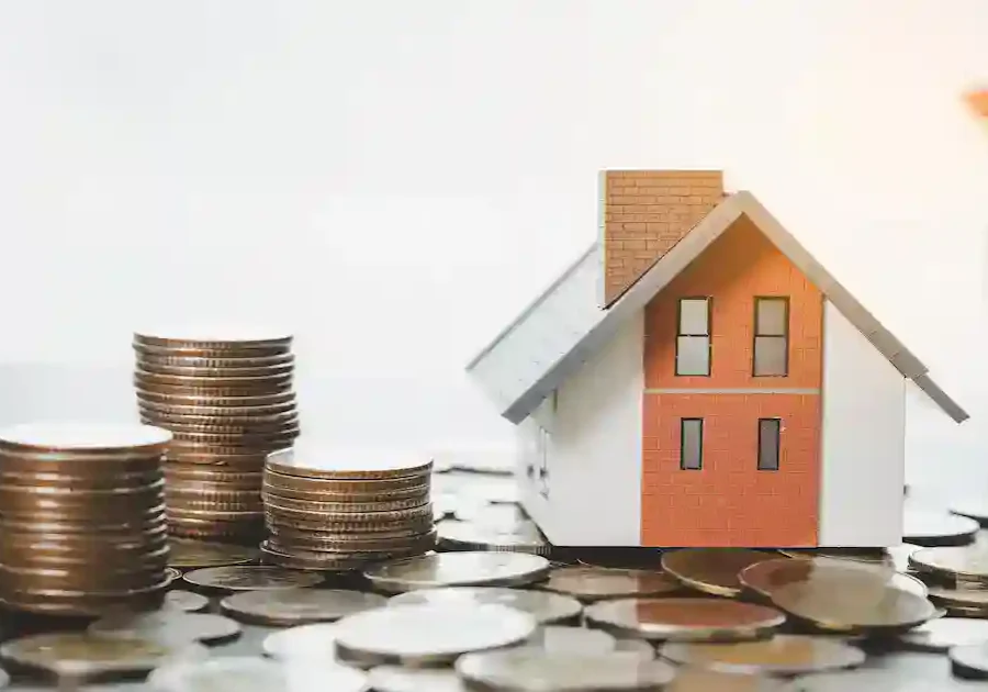 A small model house is surrounded by stacks of coins and loose coins on a table, symbolizing saving or investing money for real estate or home ownership. A blurred plant is visible in the background.