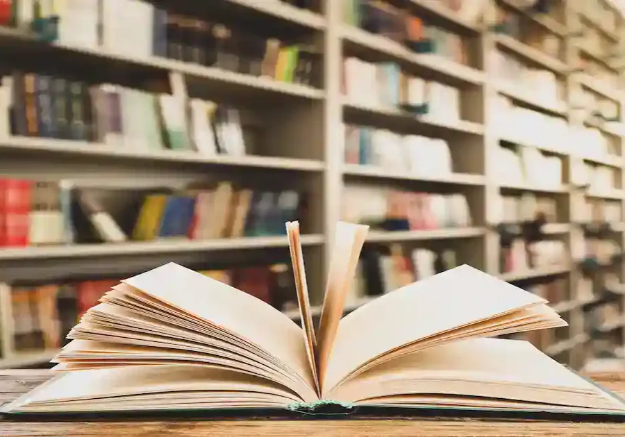 An open book sits on a wooden table in a library, with blurred bookshelves filled with books in the background.