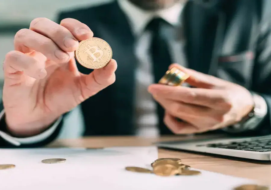 A person in a suit holding a gold Bitcoin coin near a laptop, with more coins and papers on the desk. The scene suggests a business or investment context involving cryptocurrency.