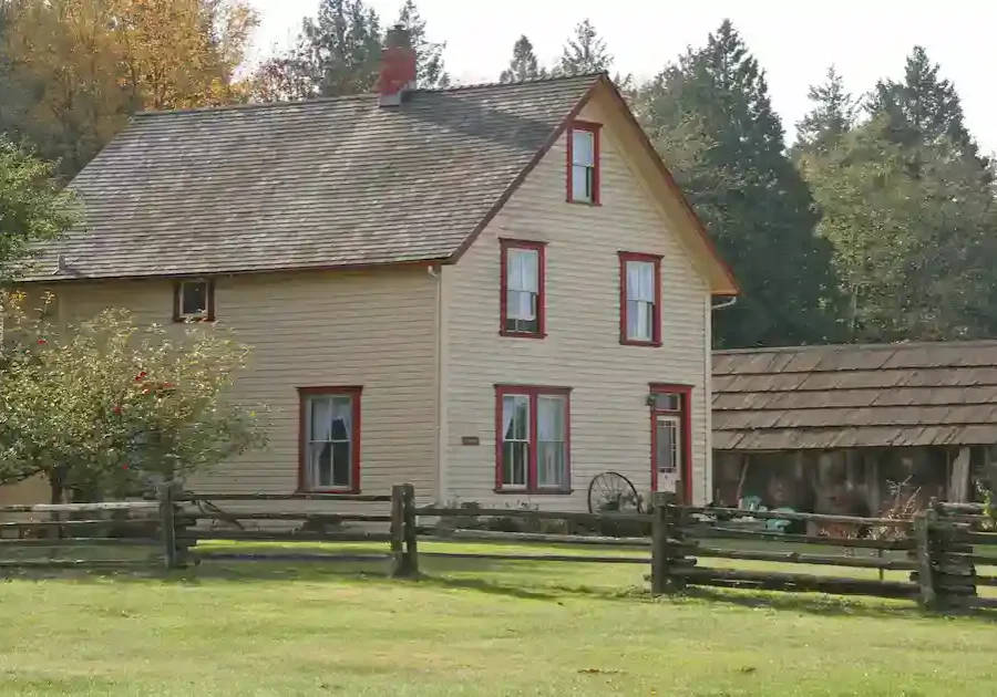 A two-story beige house with red trim sits behind a wooden fence, with a grassy yard, trees, and a rustic wooden outbuilding in the background. The scene is peaceful and surrounded by greenery.