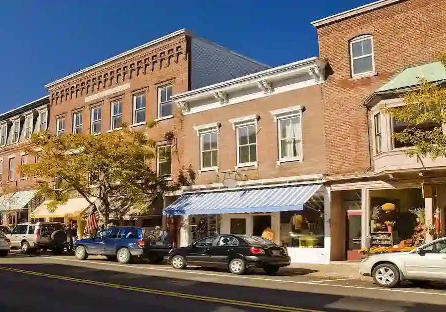 A row of brick buildings with shops and awnings along a sunny small-town street, cars parked at the curb, and trees with green leaves lining the sidewalk under a clear blue sky.